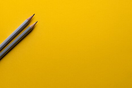 gelber Hintergrund und links ragen zwei Bleistifte ins Bild hinein.