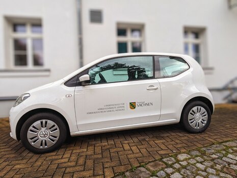 Weißes Auto mit Aufschrift der Schule steht vor einem Gebäude.